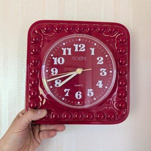 May include: A square, claret-coloured quartz clock with a white clock face. The clock face features black numerals and hands, with the word "moretime" printed in the centre. The clock has a decorative border of small, round accents.