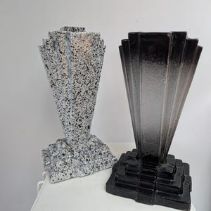 May include: Two Art Deco style vases. One is speckled with black and white, the other is black with a gradient effect. Both have a stepped base and a fan-like top. The vases are displayed on a white surface.