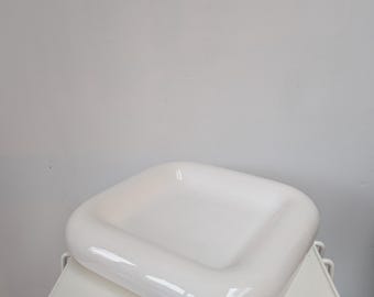Pode incluir: Um prato quadrado de cerâmica branca com cantos arredondados e um acabamento liso e brilhante. O prato apresenta uma borda larga, espessa e ligeiramente elevada que se inclina suavemente para um centro plano e rebaixado, criando uma forma.