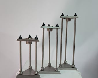 Pode incluir: Um conjunto de três castiçais de metal prateado de alturas variadas. Cada castiçal apresenta uma base triangular, três hastes de suporte verticais e um copo central circular para velas, rematado com três pequenas pontas decorativas pretas.