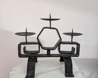 Pode incluir: Um candelabro de metal preto com acabamento texturizado e martelado. A estrutura inclui uma moldura hexagonal central ladeada por duas secções retangulares, cada uma com um prato circular e uma ponta central para segurar as velas.