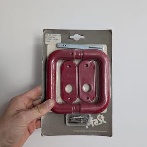 Può includere: Un set di maniglie per porte rosse, confezionato su un supporto di cartone grigio. Il set include una maniglia rettangolare, due piastre rettangolari e viti. La confezione riporta il testo "HaSt" e "PATENT ROSET HOOD".
