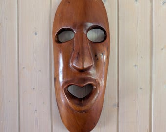 Pode incluir: Uma máscara de madeira esculpida com uma superfície lisa e polida. A máscara apresenta aberturas ovais para os olhos, um nariz proeminente e uma boca larga e aberta. A máscara é de cor castanha quente e parece estar pendurada numa parede de madeira clara.