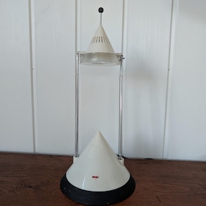 Peut inclure: Une lampe de table composée d'une base conique blanche avec un socle circulaire noir, deux tiges verticales en métal chromé et un abat-jour conique blanc réglable surmonté d'un petit embout noir avec un diffuseur translucide.