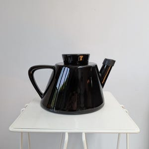 May include: A glossy black teapot with a geometric design. The teapot has a handle, a spout, and a lid. It is sitting on a white metal side table. The teapot is a modern design.