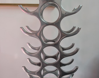 Pode incluir: Um suporte vertical para vinhos em metal prateado com acabamento polido. A estrutura apresenta uma coluna central de aberturas circulares para armazenamento de garrafas, ladeada por suportes curvos em forma de chifre em ambos os lados, montada sobre uma base quadrada.