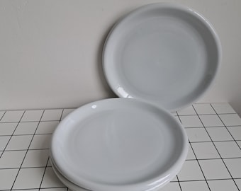 Pode incluir: Um conjunto de três pratos de cerâmica branca lisa com um acabamento suave e brilhante. Os pratos são redondos com uma borda ligeiramente elevada, apresentando uma cor branca sólida e limpa. Estão empilhados, exibindo um design uniforme.