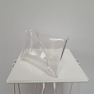 Op de afbeelding: Een heldere, transparante acrylhouder in envelopstijl met een gevouwen, geometrisch ontwerp. Het object heeft gladde, gebogen randen en een hol midden, vervaardigd uit één enkel stuk gevormd materiaal met een duidelijke diagonale vouw.
