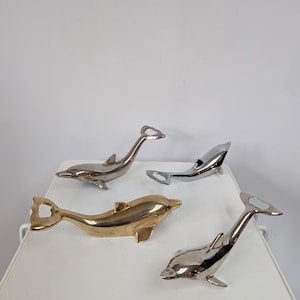 Puede incluir: Cuatro abridores de botellas con forma de delfín en tonos plateados y dorados. Los abridores están dispuestos sobre una superficie blanca, con los delfines mirando en diferentes direcciones. Los abridores son de metal y tienen un acabado brillante.