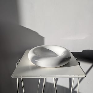 Può includere: Una ciotola in ceramica bianca, circolare e liscia, con un bordo spesso e arrotondato e un centro concavo profondo. L'oggetto presenta una finitura lucida e riflettente ed è posizionato su un supporto in metallo bianco con gambe sottili.