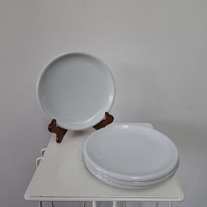 Op de afbeelding: Een set van drie effen witte keramische borden met een gladde, glanzende afwerking en een eenvoudig rond ontwerp. Eén bord staat rechtop op een donkerbruine houten standaard, terwijl de andere twee borden plat voor de standaard zijn gestapeld.