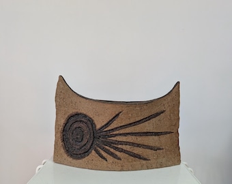 May include: A curved, tan ceramic vessel with a rough, textured surface. The front features a dark brown, circular spiral design with radiating lines extending outwards, resembling a comet. The top edge has two pointed, upward-curving corners.