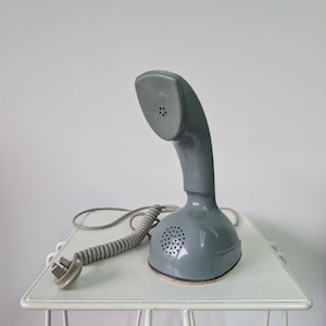 Może przedstawiać: Błyszcząca, szara, jednoczęściowa słuchawka telefoniczna o zakrzywionym kształcie z okrągłą kratką głośnika u podstawy. Szary, spiralny przewód wychodzi z boku urządzenia i kończy się standardową wtyczką elektryczną.