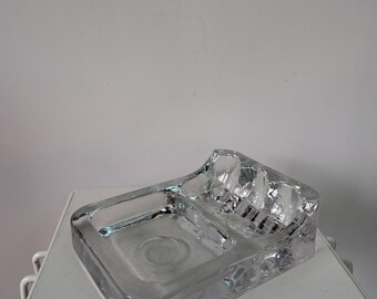 May include: A rectangular clear glass tray featuring a thick, heavy construction with a smooth, flat base section and a raised, textured section on one side that includes three distinct, rounded indentations for holding small items securely.