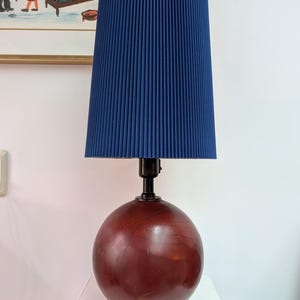 May include: A table lamp featuring a spherical, dark reddish-brown wooden base and a tall, cylindrical, pleated navy blue fabric lampshade. The lamp has a black metal socket and a simple, clean silhouette, providing a focused light source.