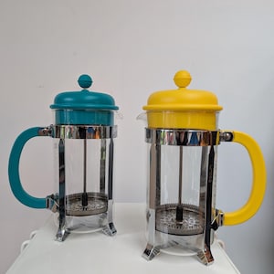 Puede incluir: Dos cafeteras de émbolo con jarras de vidrio transparente, marcos de acero inoxidable y detalles coloridos. Una tiene tapa y asa turquesa, la otra tapa y asa amarilla. Ambas están sobre una superficie blanca.