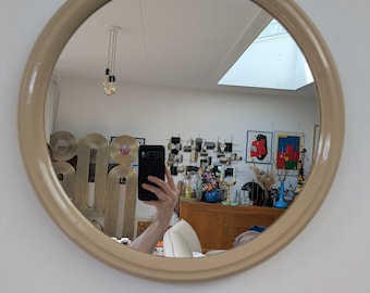May include: A circular wall mirror featuring a smooth, light beige frame. The reflective surface shows a room interior, including a wooden cabinet, various decorative objects, and a person holding a black smartphone to capture the image.