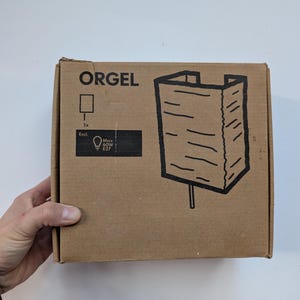 May include: A brown cardboard box with the word "ORGEL" printed on it. The box features a line drawing of a lamp and includes details about the light bulb, including a maximum of 60W and an E27 base.