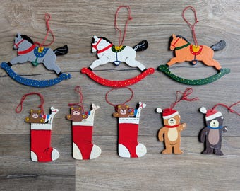 May include: A collection of nine flat wooden ornaments featuring three rocking horses in grey, white, and orange, three red stockings containing small teddy bears, and two standing teddy bears wearing red and white Santa hats, all with red string loops.