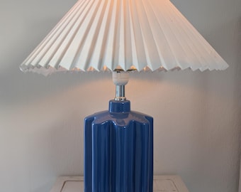May include: A blue ceramic table lamp with a white pleated lampshade. The lamp base has a starburst design.