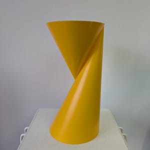 Puede incluir: Un florero de color amarillo brillante con un acabado mate suave y una forma geométrica distintiva. El objeto presenta una abertura circular ancha en la parte superior que se estrecha hacia una sección central retorcida antes de ensancharse en la base.