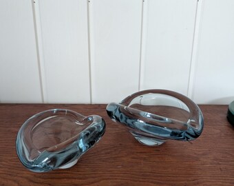 May include: Two small, clear glass bowls with a distinct blue-tinted rim and base. The bowls feature an organic, asymmetrical shape with thick, rounded edges and a smooth, polished surface, reflecting light across their curved forms.