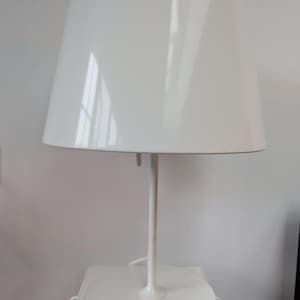 May include: A white table lamp with a conical lampshade. The lamp has a slender stem and a circular base. The lampshade is wider at the top and tapers down. The lamp is made of a glossy white material.