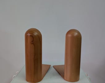 Pode incluir: Um par de suportes para livros em forma de L feitos de madeira castanho-clara com um acabamento liso e polido. Cada suporte apresenta um poste vertical cilíndrico alto com um topo arredondado em forma de cúpula e uma base plana.