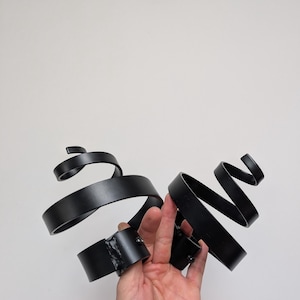 May include: Black metal sculpture with a swirling ribbon design. The sculpture has a matte finish and is held in a hand against a white background. The metal ribbons are approximately 25 cm long.