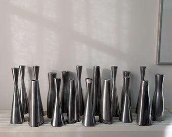 May include: A collection of seventeen silver-toned metal candle holders with a tapered, hourglass-like silhouette. Each holder features a smooth, reflective surface and a flared base, arranged in a row on a flat, light-coloured surface.