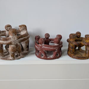 Op de afbeelding: Drie handgemaakte sculpturen die figuren tonen die elkaar omarmen. De sculpturen zijn in bruin- en roodtinten, met zichtbare textuur. De figuren zijn in een cirkel gerangschikt, met armen om elkaar heen. De sculpturen staan op een witte plank.