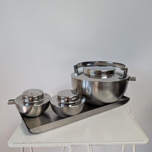 May include: A stainless steel serving set, including a large bowl with a handle, two smaller bowls with lids, and a rectangular tray. The set is displayed on a white, wire-frame table against a plain white background.