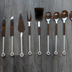 Può includere: Una collezione di utensili da cucina, tra cui un mestolo, coltelli, una paletta per torte, una forchetta, una spatola e cucchiai. Gli utensili hanno lame e teste in acciaio inossidabile con manici marroni e bianchi.