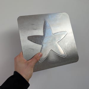 May include: A square silver-coloured metal plate featuring a centred embossed five-pointed starfish design. The surface of the metal has a brushed finish with a visible spiral pattern etched into the centre of the starfish shape.