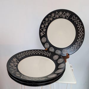 Könnte beinhalten: Ein Stapel schwarz-weißer Keramikplatten mit einem dekorativen Rand aus geometrischen Formen und Spiralen. Die Teller haben eine cremefarbene Mitte und werden auf einem weißen Metallständer präsentiert.