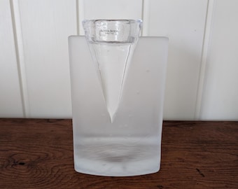 May include: A clear and frosted glass candle holder by Kosta Boda. The rectangular base features a frosted finish with a smooth, clear, cone-shaped insert at the top. The text Kosta Boda Since 1742 is printed on the clear glass rim.