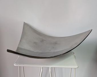 Pode incluir: Uma taça de metal prateado com formato quadrado e bordas curvas e elevadas. A taça tem uma superfície lisa e reflexiva e está posicionada sobre um suporte de metal branco com quatro pernas finas e inclinadas e um topo quadrado.