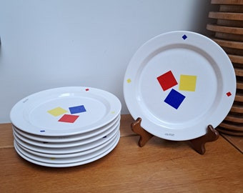 May include: A stack of white ceramic plates and one upright plate on a stand, each featuring a central cluster of red, yellow, and blue squares. The rims are decorated with small, scattered red, yellow, and blue squares, and the text "L'ARTISTE" is printed in small black letters on the rim.