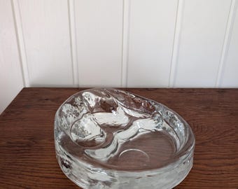 May include: A clear glass bowl with an irregular, organic shape and a thick, textured rim. The interior surface features smooth, rounded indentations, while the exterior walls have a rippled, uneven appearance that catches the light.