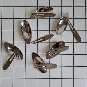 May include: A collection of polished, stainless steel serving utensils, including spoons and forks, arranged on a white tiled surface. The utensils have a sleek, modern design with elongated oval bowls and slender handles.