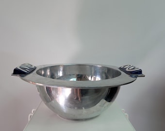 Pode incluir: Uma taça de metal prateado polido com uma borda larga e plana e duas alças decorativas. Cada alça apresenta uma base retangular azul-escura com um design gráfico abstrato de redemoinhos brancos na superfície superior da alça.