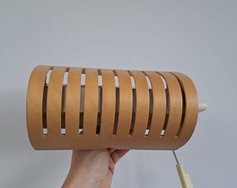 May include: A cylindrical light fitting featuring a light brown wood veneer exterior with vertical slats that allow light to pass through. The lamp includes a white power cord with an integrated plastic switch attached to the side of the housing.