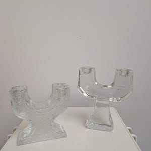 Könnte beinhalten: Zwei klare Kerzenhalter aus Glas mit einem einzigartigen, geschwungenen Design. Jeder Halter hat zwei Öffnungen für Kerzen und einen stabilen Sockel. Das Glas hat eine strukturierte Oberfläche, die dem schlichten, eleganten Design visuelles Interesse verleiht.