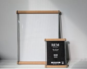 May include: Two rectangular poster hangers with light wood dowels and thin metal wire frames. The smaller hanger holds a black card with white text reading REM, 10 x 15 cm, 3 7/8 x 5 7/8 in, and the IKEA logo. The larger hanger is empty with a clear panel.