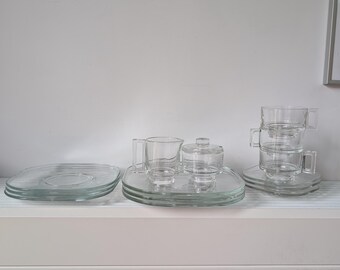 May include: A clear glass tea set consisting of a stack of square saucers, a creamer pitcher, a lidded sugar bowl on a square tray, and a stack of three glass cups with square handles, all featuring a smooth, transparent finish.
