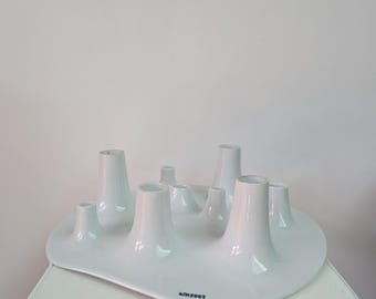 Pode incluir: Um castiçal de cerâmica branca com uma forma orgânica e fluida, apresentando múltiplas aberturas cilíndricas elevadas de alturas variadas. A base é lisa e assimétrica, com o texto preto "WJ 2003" impresso perto da borda frontal.
