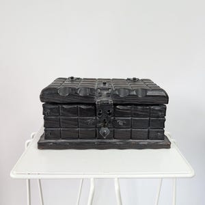 Puede incluir: Un cofre de madera oscura con forma rectangular y tapa con bisagras. El cofre presenta un patrón de rejilla y herrajes metálicos, incluyendo un pestillo y detalles decorativos. El cofre se exhibe sobre una mesa de estructura metálica blanca.