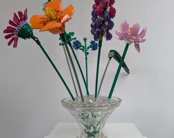 May include: A bouquet of colourful plastic building block flowers in a clear, faceted glass vase. The arrangement includes an orange flower, a purple and pink lupine-style stalk, a pink lotus-like bloom, and small blue accents on green stems.