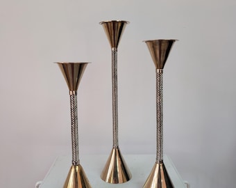 Pode incluir: Três castiçais metálicos de alturas variadas, cada um com uma base e topo cónicos em tom dourado polido, ligados por um caule fino prateado com um design de corda torcida. O design é uniforme em todas as três peças verticais.
