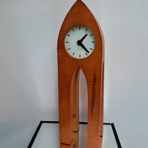 Puede incluir: Un reloj de madera con una esfera clara y manecillas negras. El reloj está alojado en un marco de madera alto y arqueado. La esfera del reloj tiene marcas negras y la marca "Karlsson". El reloj es un elemento decorativo para el hogar.